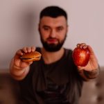 A man holds an apple and a cheeseburger, showcasing a choice between healthy and unhealthy food.