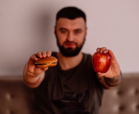 A man holds an apple and a cheeseburger, showcasing a choice between healthy and unhealthy food.
