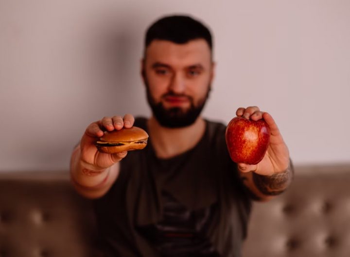 A man holds an apple and a cheeseburger, showcasing a choice between healthy and unhealthy food.