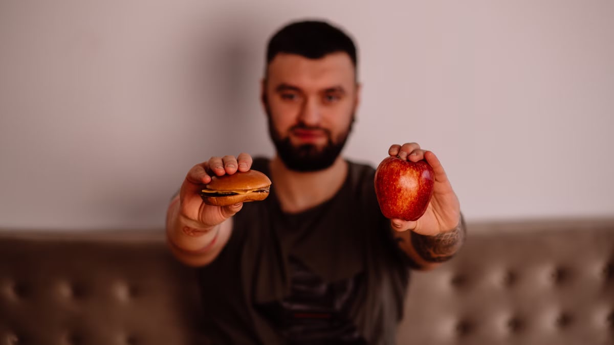 A man holds an apple and a cheeseburger, showcasing a choice between healthy and unhealthy food.